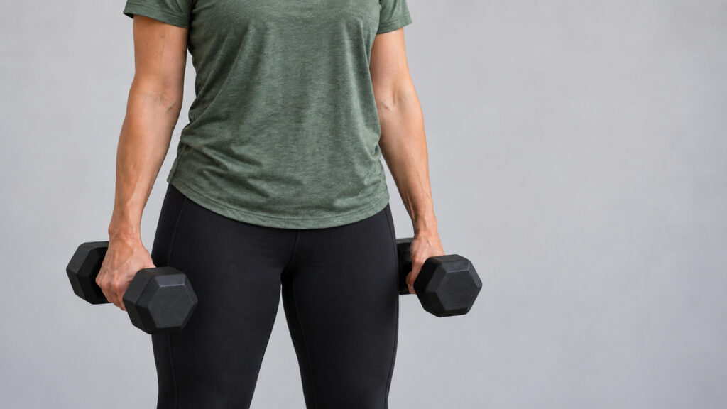 Vrouw in een groen sportshirt en zwarte sportlegging houdt twee dumbbells vast.